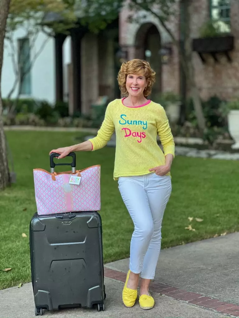 travel look for woman over 50 standing next to luggage wearing a yellow sunny days sweater and white pants with yellow shoes