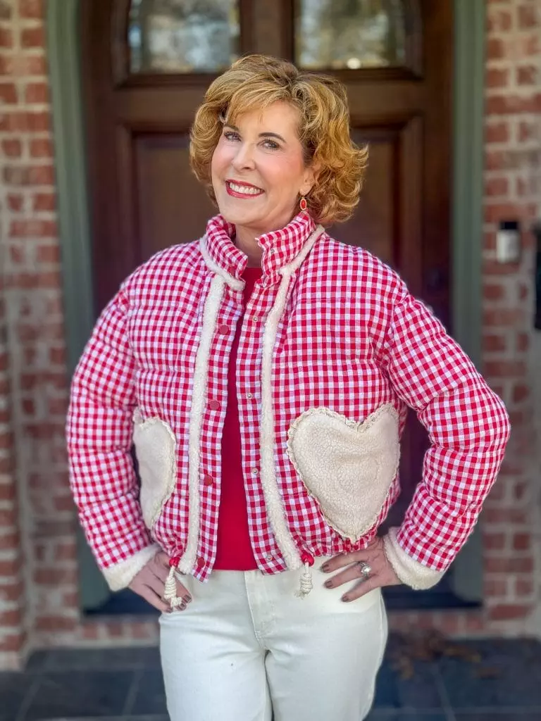 Woman standing outside in a red gingham jacket with heart shaped sleeves and cream colored jeans and a red cashmere sweater underneath the jacket